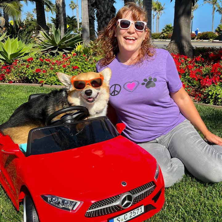 Dog mom wearing a purple t-shirt with Peace, Love and Paw Symbols sitting next to a corgi in a red mercedes toy car.