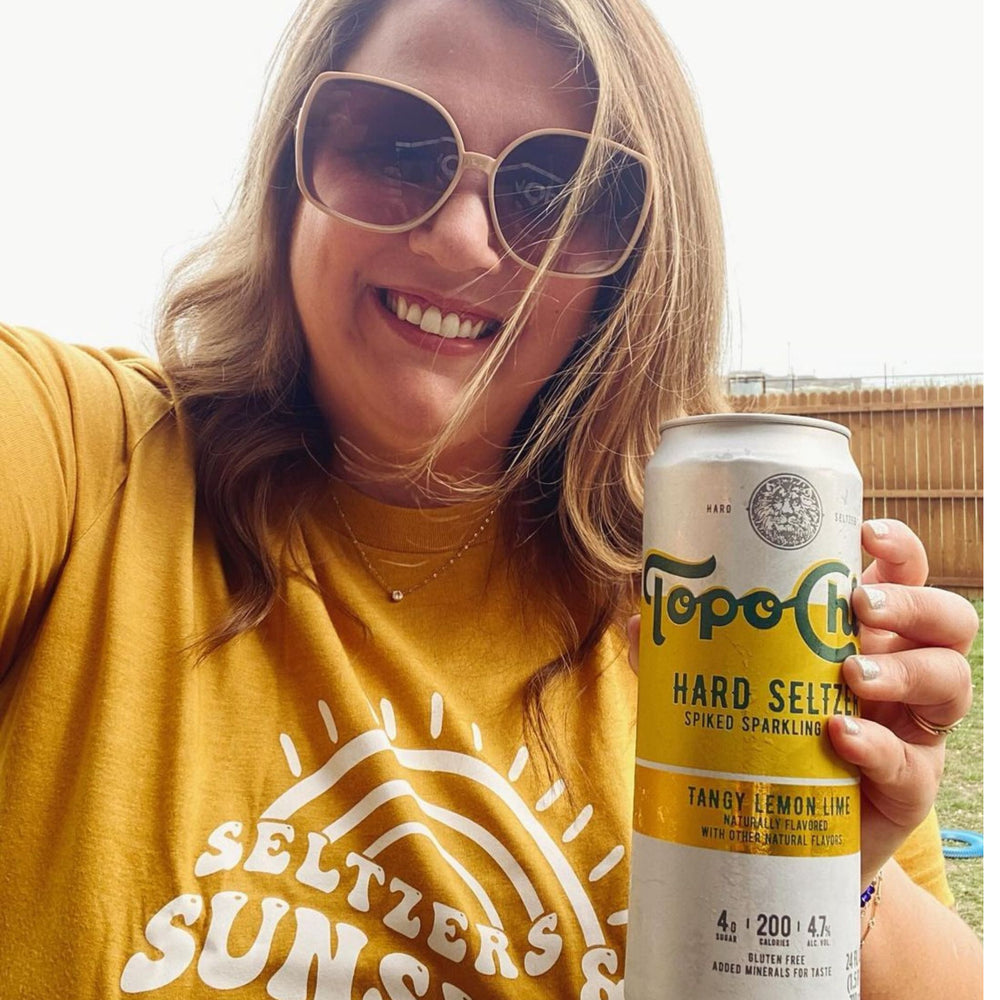 Female wearing a graphic shirt that says seltzers and sunshine on gold color t-shirt. She is holding a seltzer drink in her hand.